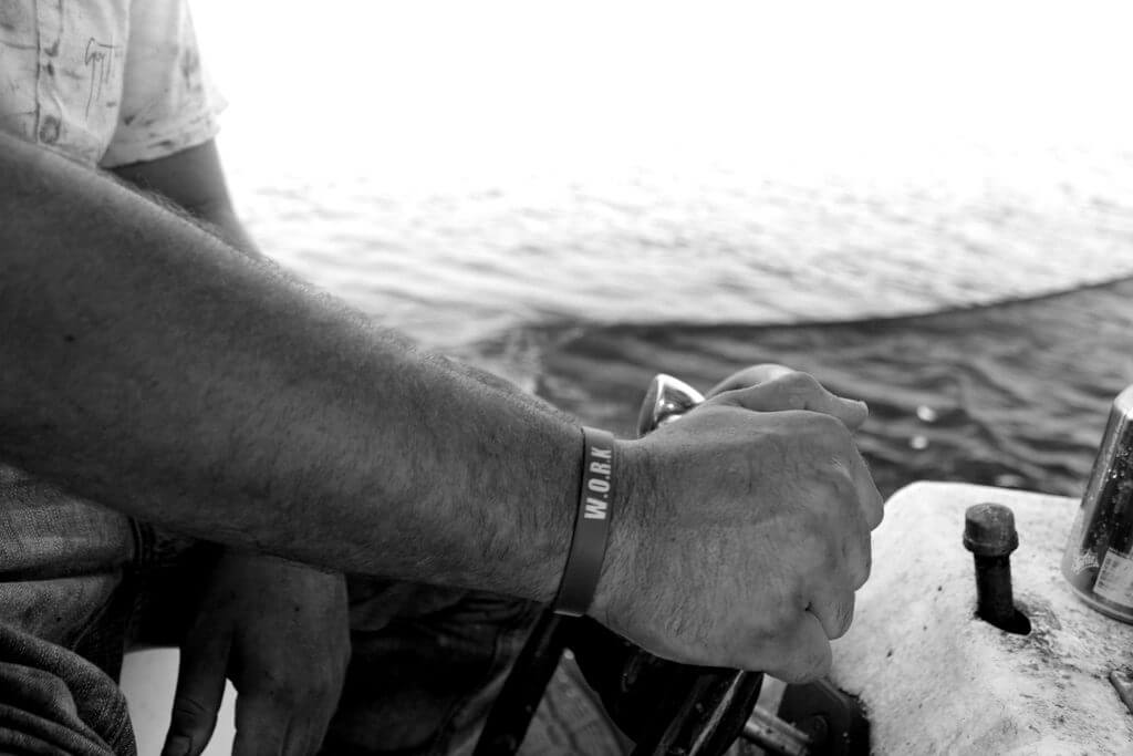 Two hands grip a boats steering wheel. The wrist closest to the camera wears a bracelet that reads "W.O.R.K."