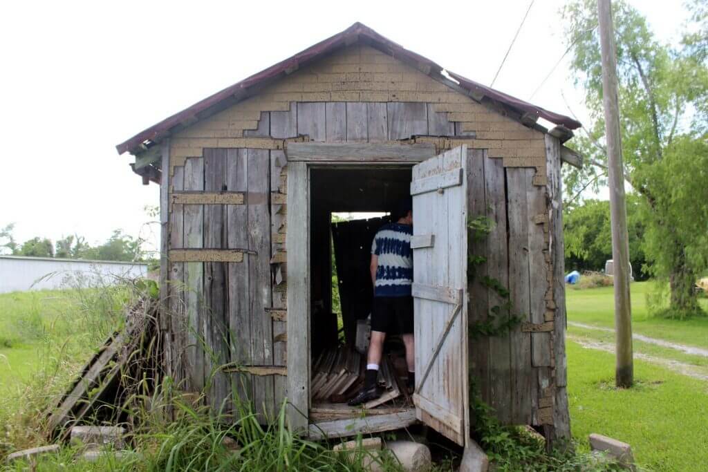 A man examines the inside of an old, dilapidated, wooden shed next to a body of water.