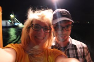 A blond, light skinned woman in glasses smiles as she takes a selfie of herself and a light skinned man in glasses and a hat. The background is dark and water can be seen behind them. 