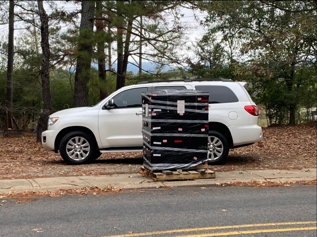 The monument, wrapped in black packing materials, arrives at the cemetery and sits on a pallet next to a medium sized SUV.