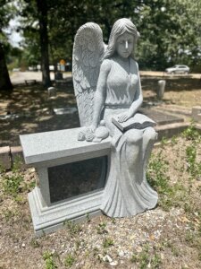 A gray granite cemetery monument depicting a seated angel with folded wings, long hair, and a flowing dress, holding an open book on her lap. She sits beside a rectangular granite bench, with carved roses at her side. The monument stands in a cemetery beneath trees, with gravestones and a road visible in the background.