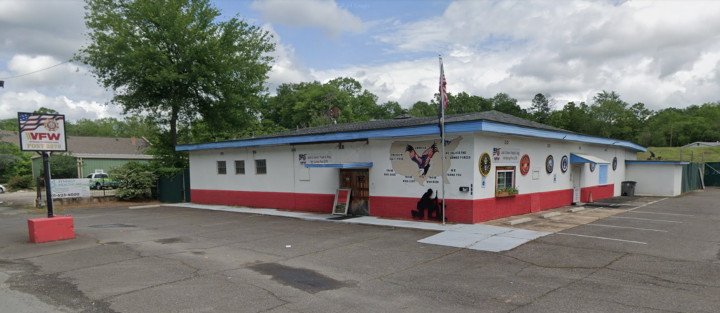 A single story building surrounded by an empty parking lot. Formerly a gay bar, the building is now a VFW and is prominently covered in military and patriotic symbols.