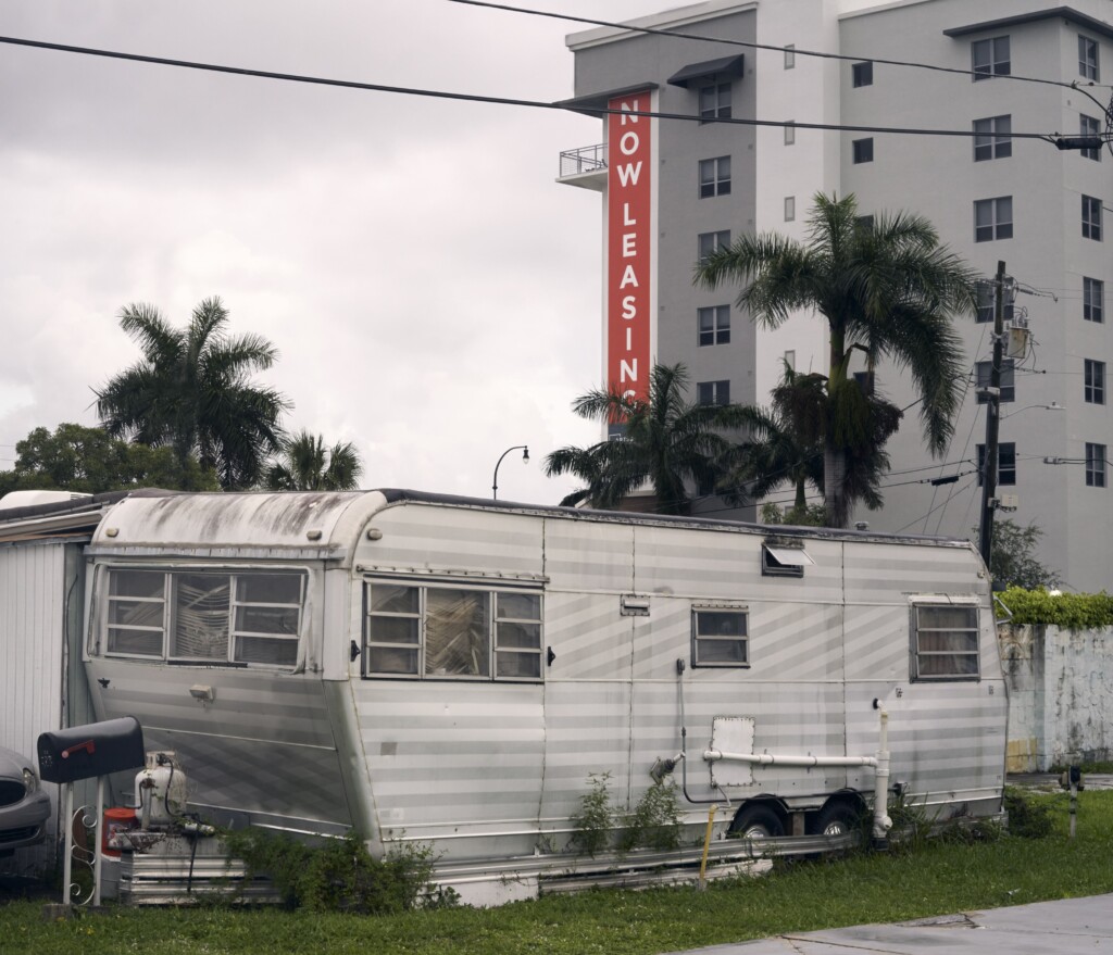 A “Now Leasing” sign on a new high rise building jabbing like a dagger into the belly of a mobile home.