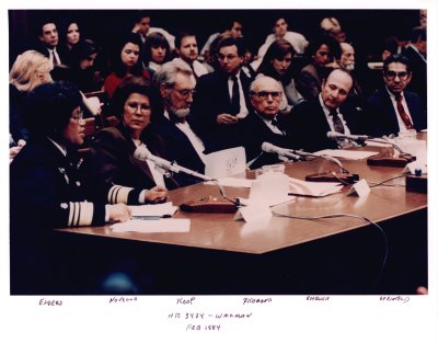 Dr. Joycelyn Elders speaks into a microphone at a Congressional hearing. She sits next to other former Surgeons General, who are listening to her with their heads turned towards her.